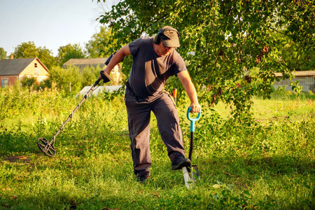 A man uses a metal detector while kneeling in tall grass, searching for treasures in a field at...