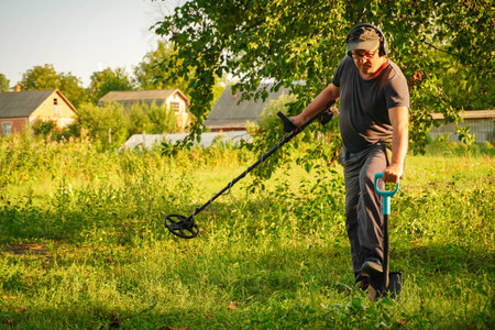 A man uses a metal detector in a vibrant green field surrounded by trees and rustic structures. The summer sun casts a warm glow as he carefully scans the ground, focused on finding hidden treasures.の写真素材