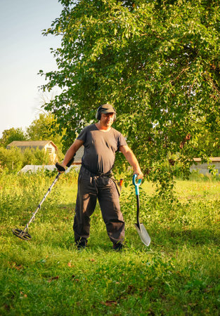 A man wearing a cap stands in a grassy field, holding a metal detector in one hand and a shovel in...
