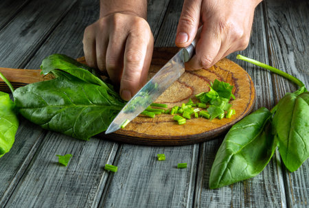 Hands chop fresh spinach leaves on a wooden surface, creating small pieces for a healthy meal. The natural light highlights the fresh greens and rustic setting.の写真素材