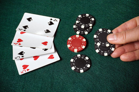 A hand holds a black poker chip, and on the green table cloth lie four playing cards with the image...