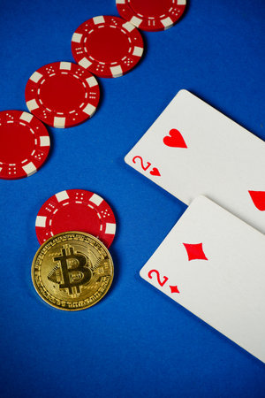 On a blue table, red poker chips are arranged alongside two playing cards. A Bitcoin coin is placed...