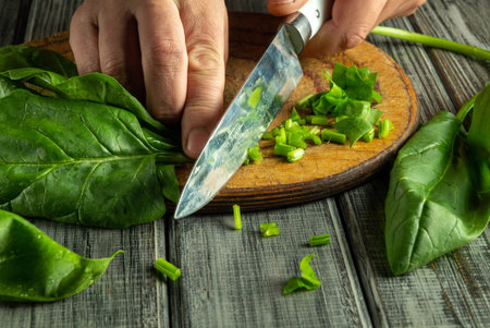 Hands are carefully cutting fresh green leaves on a wooden board, with some chopped greens scattered around. The light creates a warm atmosphere in the kitchen.の写真素材