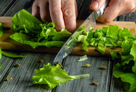 Hands skillfully chop fresh lettuce on a wooden cutting board in a bright kitchen. Colorful greens are spread around, showcasing a focus on healthy cooking.の写真素材