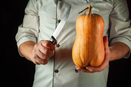 A chef wearing a white coat holds a butternut squash in one hand and a knife in the other, ready to start preparing a meal. The background is dark, emphasizing the focus on the chef and ingredients.の写真素材