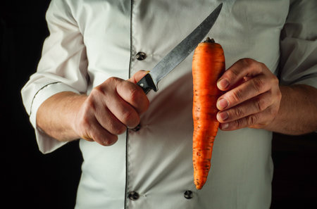 A chef holds a bright orange carrot in one hand while using a sharp knife in the other hand,...