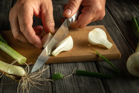 Hands expertly slice fresh green onions on a wooden board. The setting features a rustic kitchen with natural light illuminating the scene, capturing a moment of cooking preparation.の写真素材
