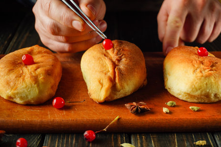 Skilled hands use tweezers to position bright cherries on warm, golden pastries. The cozy kitchen is filled with spices and a rustic wooden board enhances the scene.の写真素材