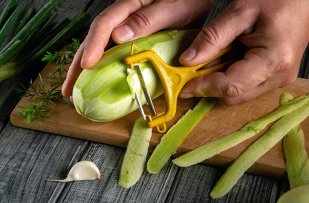 Hands work diligently to peel a fresh zucchini using a yellow peeler. Fresh herbs and a clove of garlic hint at plans for a delicious meal in a cozy kitchen.の写真素材