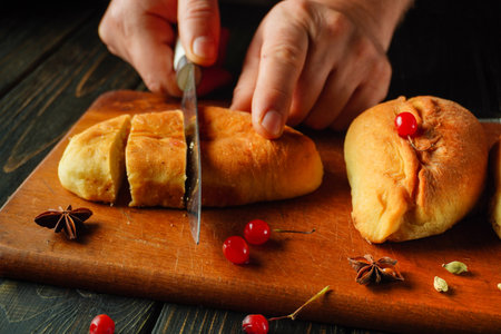 Hands delicately slice warm bread on a wooden board. Vibrant cherries and spices surround the bread, creating a warm and inviting atmosphere in the kitchen.の写真素材
