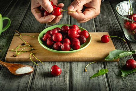Hands remove stems from ripe cherries while a green bowl filled with the fruit sits on a wooden...