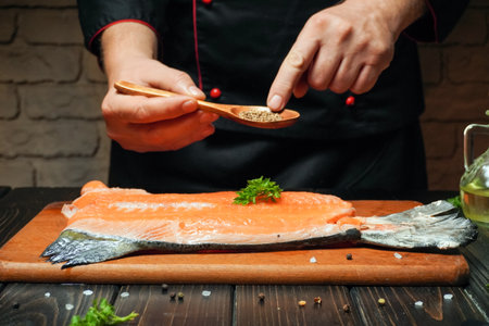 A chef carefully sprinkles spices over a fresh salmon fillet placed on a wooden cutting board. The kitchen features warm lighting and rustic brick walls.の写真素材