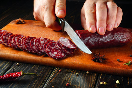 Hands carefully slice a piece of cured sausage into even rounds on a wooden cutting board, surrounded by colorful spices and herbs in a cozy kitchen setting.の写真素材