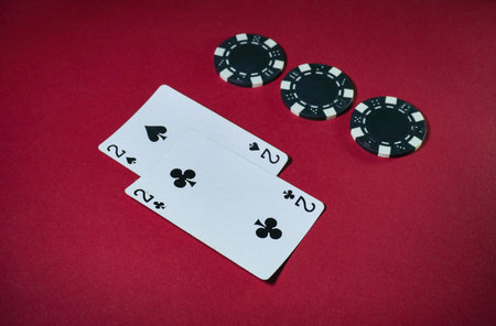 Two playing cards, two of spades and two of clubs, are placed on a red table alongside several black poker chips, hinting at a competitive game night atmosphere.の写真素材