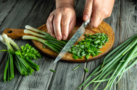 Hands are skillfully chopping fresh green onions on a wooden cutting board. The scene shows vibrant vegetables and kitchen tools, highlighting the act of preparing ingredients for cooking.の写真素材