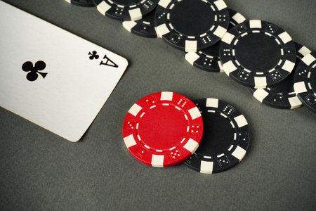 A red poker chip and black chips surrounding an ace of clubs on a gray table surface indicate a thrilling card game. Players are focused, enjoying their time.の写真素材