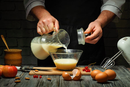 A chef pours milk from a glass jug into a bowl containing eggs and other ingredients. The kitchen is dimly lit, creating a warm ambiance as fresh apples and eggs are nearby.の写真素材