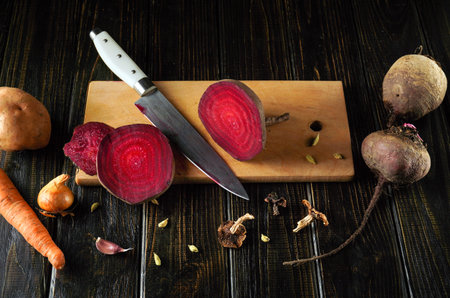 Freshly cut red beets and various vegetables are displayed on a wooden cutting board. A sharp knife and scattered ingredients create an inviting scene for cooking.の写真素材