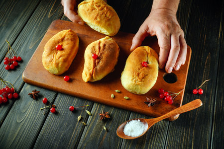 Hands carefully arrange freshly baked bread rolls topped with cherries, surrounded by spices and salt on a wooden cutting board, showing a delightful culinary moment.の写真素材