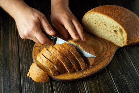 Hands cut soft, golden-brown bread into even slices on a rustic wooden cutting board. The warm...