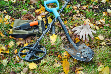 A metal detector, shovel, and gloves rest on the ground surrounded by colorful autumn leaves. Several coins are visible, showcasing a successful treasure hunt in a park.の写真素材