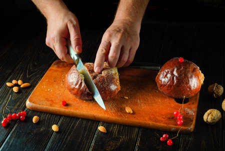 Freshly baked bread is being sliced on a wooden board by hands holding a sharp knife. Nuts and...