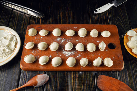 Freshly made dumplings rest on a wooden cutting board. Flour is strewn about, and utensils sit nearby, showcasing a hands-on cooking process in a warm kitchen environment.の写真素材