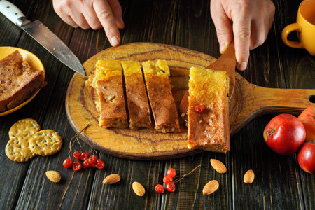 A person carefully slices a homemade cake on a wooden board. Nearby, fresh apples and nuts complement the dessert. The scene is warm and inviting.の写真素材