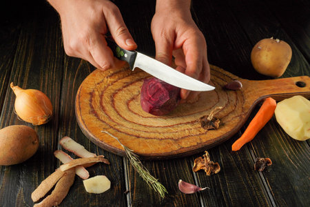 A chef carefully slices a red beet on a wooden cutting board. Surrounding them are various...