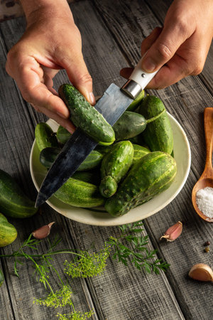 Fresh cucumbers are being carefully sliced by hands holding a knife over a bowl in a rustic kitchen. Garlic and salt are nearby, adding to the culinary atmosphere.の写真素材