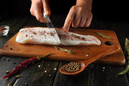 A person cuts into a fresh fish fillet placed on a wooden cutting board. Various spices and herbs...