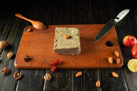 A wooden cutting board displays a square block of nut-based dessert topped with an almond. Fresh fruits and spices surround it, showcasing the ingredients used for preparation.の写真素材