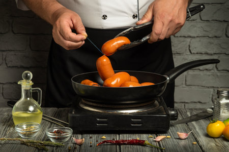 A cook holds tongs while preparing sausages in a frying pan on a stove. Ingredients like oil and seasonings are placed on the table for the cooking process.の写真素材