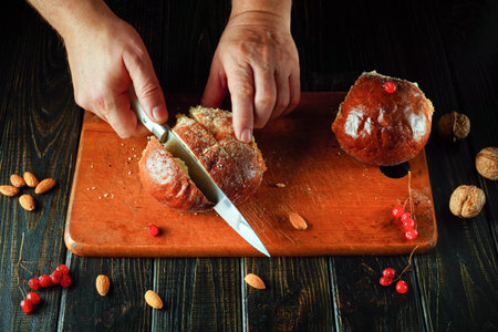 Two hands skillfully slice a round loaf of bread on a wooden cutting board. Surrounding the bread are nuts and red berries, adding color to the kitchen scene during the day.の写真素材