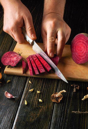 Hands are skillfully cutting vibrant red beets into slices on a wooden surface. Various spices and garlic are nearby, reflecting a cozy cooking environment.の写真素材