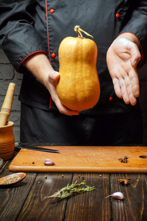 A chef in a dark jacket holds a ripe butternut squash above a wooden cutting board, surrounded by fresh herbs and spices in a kitchen filled with warm, inviting light.の写真素材
