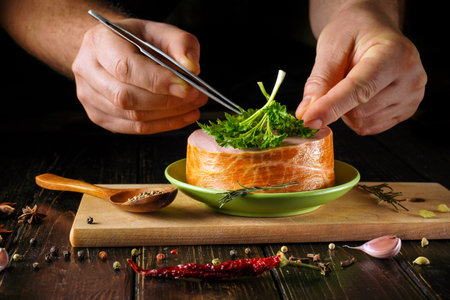 A chef uses tweezers to place fresh herbs on a plate with meat on a wooden surface. Spices and garlic surround the plate, adding to the kitchen atmosphere in the late evening.の写真素材