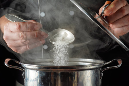 A chef pours salt from a spoon into a pot on the stove. Steam rises from the pot as the chef adds...