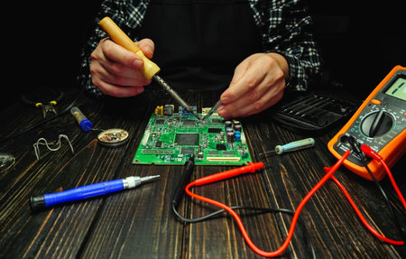 A man works on a circuit board at a wooden table with various tools and a multimeter. The setting is...