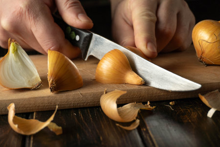 Hands chop onions on a wooden cutting board using a knife. Peels and chopped pieces are scattered...