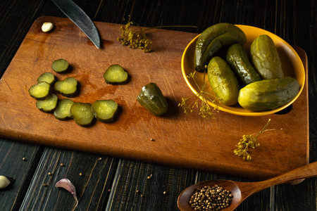 A wooden board holds sliced pickles and whole green pickles in a yellow bowl. A knife is placed nearby, and spices scatter around the surface. The scene shows kitchen preparation.の写真素材