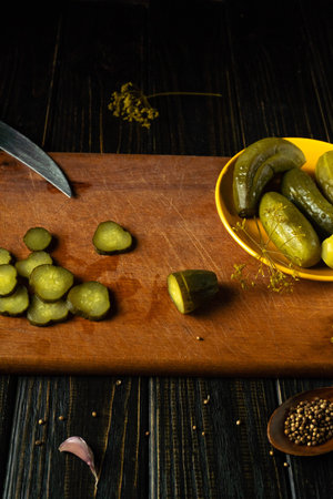 Sliced pickles lie on a wooden board next to whole pickles in a yellow bowl. A knife is placed nearby as garlic and mustard seeds are scattered on the surface.の写真素材