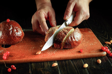 Hands hold a knife to slice bread on a wooden cutting board. Red berries and almonds are nearby....