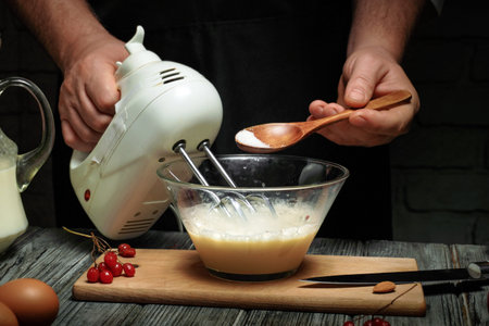 A person is mixing batter using a hand mixer in a clear bowl. They are adding sugar with a wooden...