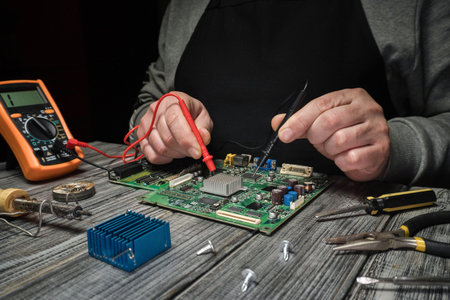 A person holds a multimeter and tests a circuit board in a workshop. Tools and small parts are...