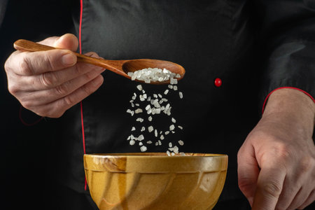 A chef holds a wooden spoon filled with coarse salt over a wooden bowl in the kitchen. The chef is focused on adding salt. This activity is part of cooking.の写真素材