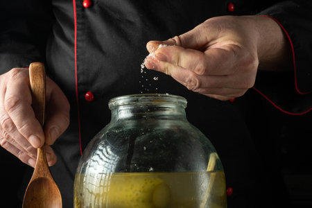 A chef prepares pickles in a kitchen by adding salt to a large glass jar. The chef uses a wooden...