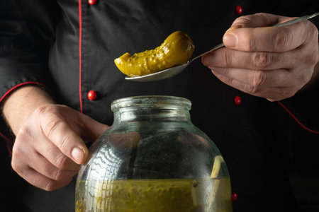 A chef is holding a spoon with a pickle above a large jar filled with pickles. The cook wears a...