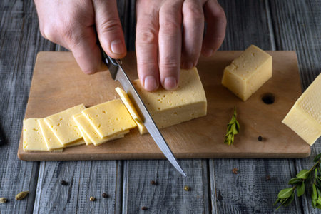 A person uses a knife to cut cheese into slices on a wooden cutting board. Fresh herbs and spices...