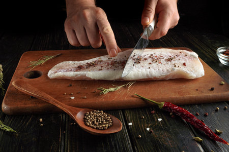 A person is cutting a fish fillet on a wooden cutting board. Spices like black pepper and dried...